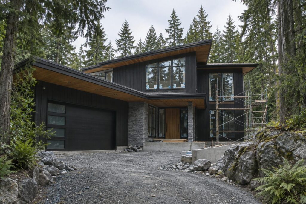 Newly built West Coast style home with dark siding and cedar soffits nearing completion on a treed lot