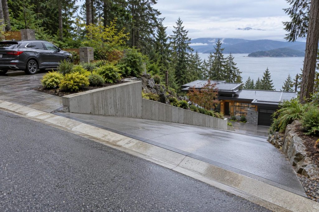 Steep paved driveway dropping from a quiet street down to a house built below road level, with a low retaining wall along the edge