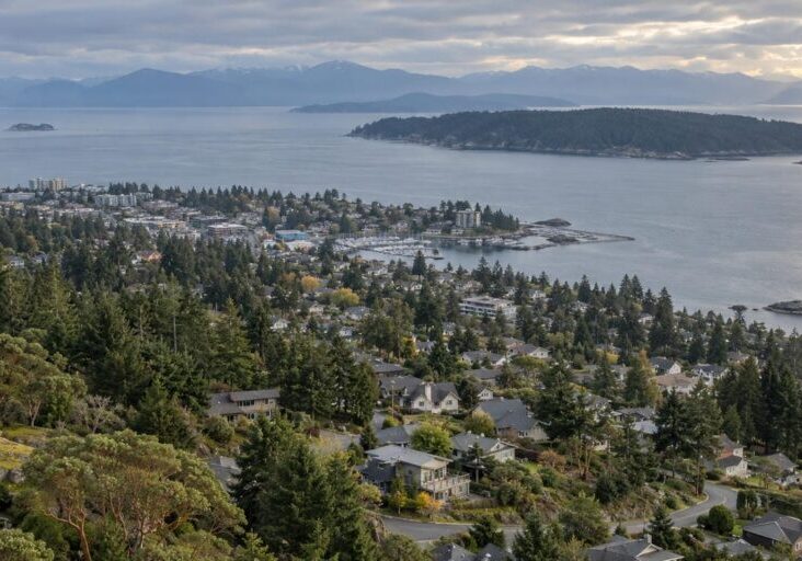 Aerial view of forested Nanaimo neighbourhoods stepping down a gentle slope toward the Strait of Georgia, with a wooded island and coastal mountains across the water.